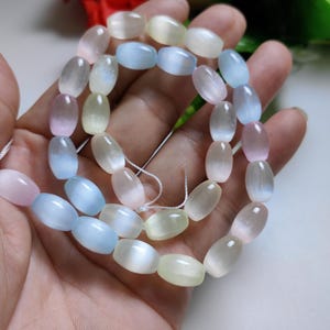Puede incluir: Una hebra de cuentas de piedras preciosas ovaladas en colores pastel rosa, azul y amarillo. Las cuentas tienen un acabado brillante e iridiscente.