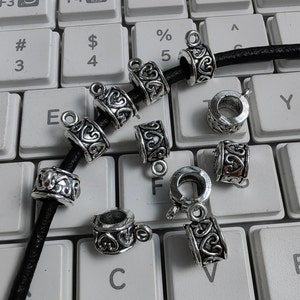 Könnte beinhalten: Silberne Metallperlenkappen mit Herzdesign. Die Perlenkappen liegen auf einer Tastatur und einem schwarzen Lederband.
