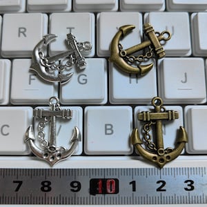 Könnte beinhalten: Fünf silberne und bronzene Anker-Anhänger auf einer weißen Tastatur mit einem Lineal, das 10 cm misst. Die Anker sind in verschiedenen Größen und Stilen.