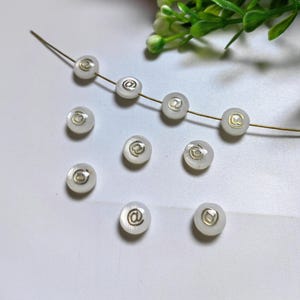 Könnte beinhalten: Eine Schnur aus weißen runden Perlen mit einem goldenen @-Symbol auf jeder Perle.