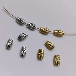 Puede incluir: Una colección de cuentas de amuleto de cigarra plateadas y doradas. Las cuentas están enhebradas en un alambre dorado. Las cuentas son de diferentes tamaños y formas.