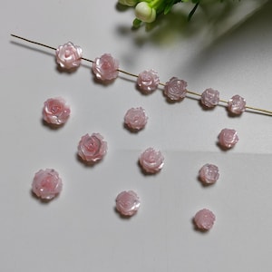 May include: A collection of pink shell rose flower beads. The beads are arranged in a line and scattered on a white surface.