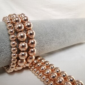 May include: Several rose gold beaded bracelets are displayed. The bracelets are made of round, shiny beads in varying sizes. The bracelets are stacked on a gray, cylindrical surface, with some spilling over onto a white surface.