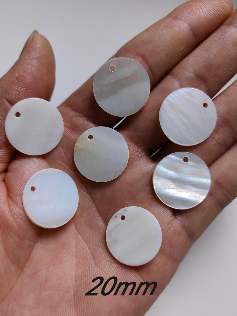 May include: Six white, round, iridescent mother of pearl shell discs with a hole in the center. The discs are 20mm in diameter and are arranged in a semi-circle on a hand.