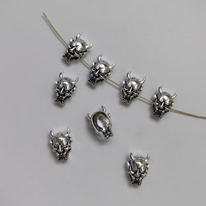 May include: Silver-toned metal charms shaped like Japanese demon masks with horns. The charms are strung on a thin wire.