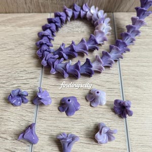 May include: A string of gradient purple flower-shaped beads arranged in a circular pattern, accompanied by several loose individual flower beads. The text G# Gradient Purple and findingscity are visible. The beads feature a smooth, glossy finish.