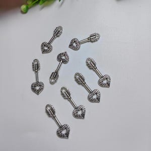 May include: Silver arrow charms with heart-shaped tips and feathered fletching. These small charms are suitable for jewellery making or craft projects. The charms are scattered on a white surface.