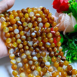 Può includere: Una collana di perle di agata gialle e bianche. Le perle sono rotonde e hanno una finitura liscia e lucida. Le perle sono infilate su un filo elastico trasparente.