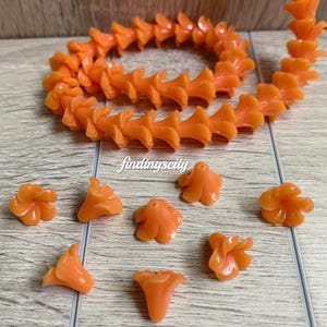 May include: A strand of orange flower-shaped beads and several loose matching beads. The text J# Orange and findingscity are visible. The beads have a smooth, glossy finish and a central hole for stringing, arranged in a circular pattern.