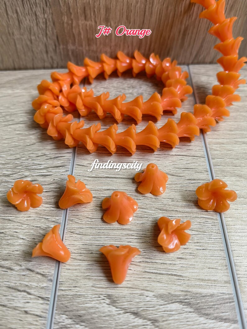 May include: A strand of orange flower-shaped beads and several loose matching beads. The text J# Orange and findingscity are visible. The beads have a smooth, glossy finish and a central hole for stringing, arranged in a circular pattern.