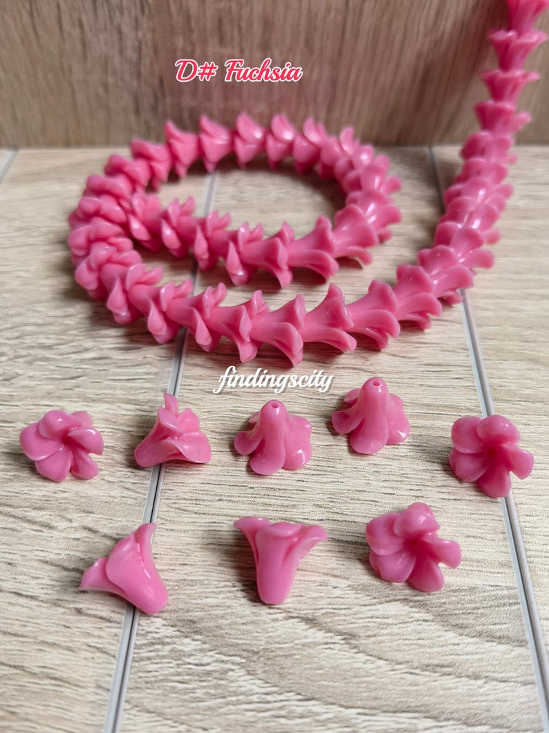 May include: A long strand of fuchsia-colored flower-shaped beads arranged in a coil, accompanied by several loose individual flower beads. The text D# Fuchsia and findingscity are visible. Each bead features a central hole for stringing.