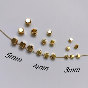 Puede incluir: Cuentas de metal de color dorado en tres tamaños: 3 mm, 4 mm y 5 mm. Las cuentas son de forma cuadrada con un agujero en el centro. Las cuentas están dispuestas en línea sobre una superficie blanca.