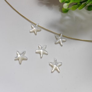 Pode incluir: Cinco pequenas contas brancas peroladas em forma de estrela-do-mar. As contas estão dispostas em um fio fino dourado, com algumas espalhadas em uma superfície branca. As estrelas-do-mar têm um brilho sutil e iridescente.
