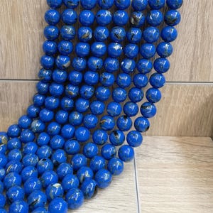 May include: A collection of round, polished beads in a vibrant royal blue color. Each bead features subtle, dark, and metallic-toned inclusions that create a marbled effect across the smooth, spherical surface of the individual jewelry components.