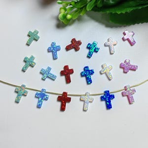 Puede incluir: Una colección de pequeñas cuentas en forma de cruz, coloridas. Las cruces son de varios colores, incluyendo azul, rojo, verde y morado, con un acabado iridiscente, similar al ópalo. Algunas están ensartadas en un fino alambre dorado, para joyería.