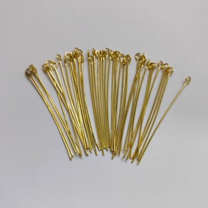 Puede incluir: Un grupo de alfileres de cabeza dorados con un bucle en la parte superior. Los alfileres están dispuestos en forma de abanico sobre un fondo blanco.