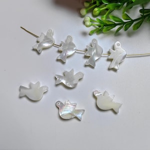 Könnte beinhalten: Ein Set aus sieben weißen Perlmutt-Taubenanhängern mit irisierendem Schimmer. Die Tauben sind auf einem goldenen Draht aufgefädelt und perfekt für die Schmuckherstellung.