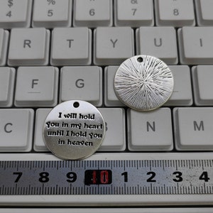 May include: Two silver metal charms. One charm is round and has a textured surface. The other charm is round and has the text "I will hold you in my heart until I hold you in heaven".