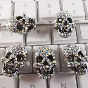 Peut inclure: Cinq perles en forme de crâne argentées avec des strass irisés. Les perles sont disposées sur un clavier.
