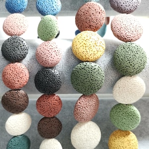 Puede incluir: Una variedad de cuentas redondas de roca volcánica en diferentes colores, incluyendo negro, marrón, blanco, verde, rojo y amarillo. Las cuentas están dispuestas en filas sobre una superficie blanca.