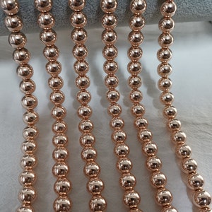 May include: Close-up of several strands of rose gold beaded jewelry. The beads are round and evenly spaced, reflecting light. The jewelry is likely a necklace or bracelet, showcasing a simple, elegant design.