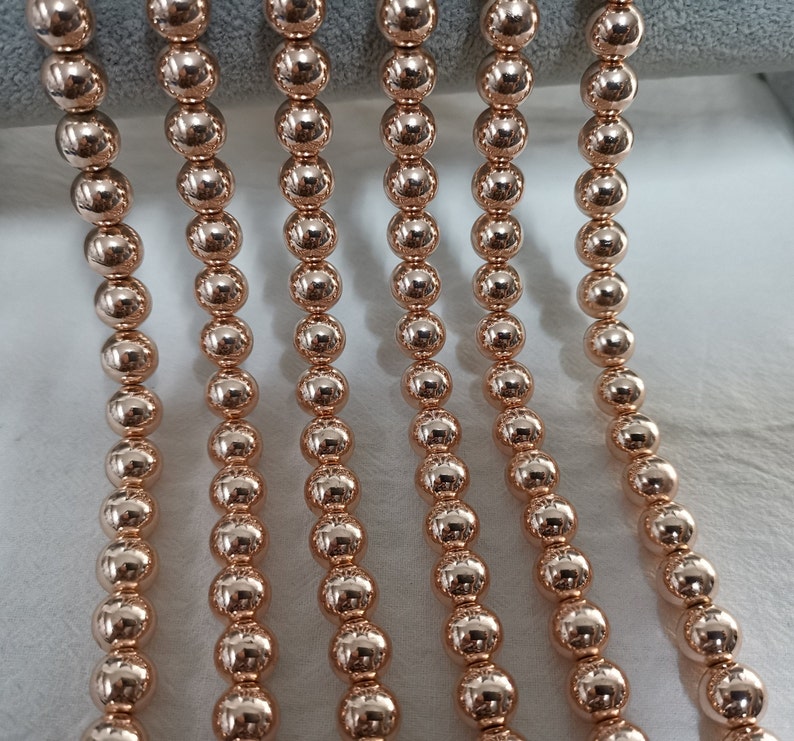 May include: Close-up of several strands of rose gold beaded jewelry. The beads are round and evenly spaced, reflecting light. The jewelry is likely a necklace or bracelet, showcasing a simple, elegant design.