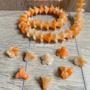 May include: A strand of gradient orange flower-shaped beads arranged in a coil, accompanied by several loose individual flower beads. The text F# Gradient Orange and findingscity are visible on the image. The beads feature a translucent finish.