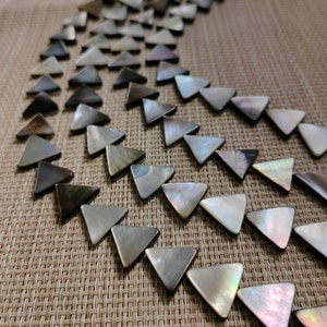 May include: A close-up of a curved line of iridescent, triangular, mother-of-pearl shell beads. The beads are arranged in a semi-circle pattern on a woven, beige surface.