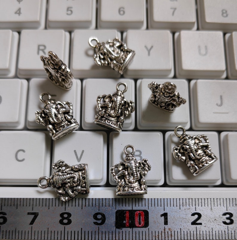 May include: A collection of silver-colored Ganesha charms, each with a loop for hanging. The charms are intricately detailed and displayed on a white keyboard. A ruler is at the bottom, showing measurements.