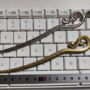 May include: Two decorative hair sticks. One is silver-coloured, the other is bronze. Both have a curved, cloud-like design at the top and a textured, elongated body. A ruler shows the sticks are about 18 cm long.