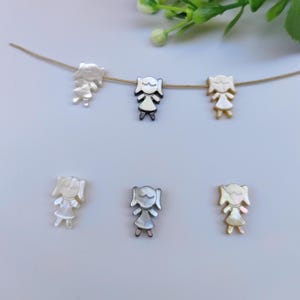 May include: Six small, girl-shaped charms in various colors, including silver, gold, and mother-of-pearl. The charms are strung on a thin, gold-colored wire and are arranged against a light background. The charms have a simple, stylized design.