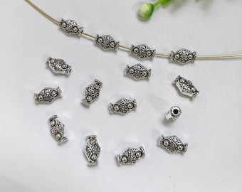 May include: Silver metal beads with a decorative design. The beads are oval shaped and have a floral pattern.
