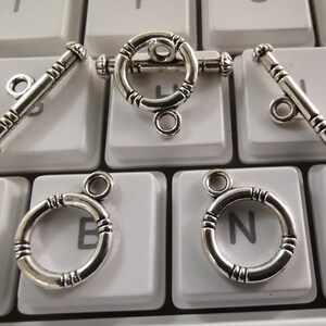 Könnte beinhalten: Fünf silberfarbene Metallverschlüsse zum Schmuckmachen. Zwei sind Toggle-Verschlüsse mit einer Stange und einem Ring. Drei sind runde Verschlüsse mit einem Ring.