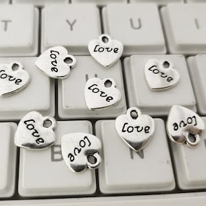Op de afbeelding: Een verzameling zilverkleurige hartvormige bedels, elk met het woord "Love" in een sierlijk lettertype. De bedels liggen verspreid op een wit toetsenbord, wat een romantisch thema creëert.