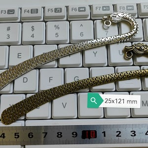 Könnte beinhalten: Zwei Drachen-Lesezeichen, eines in Silber und eines in Gold, mit einem schuppigen Design. Die Lesezeichen sind 25 x 121 Millimeter groß.