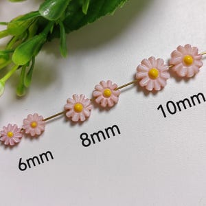 Puede incluir: Un conjunto de cinco cuentas de flores de margarita rosas con centros amarillos enhebradas en un alambre dorado. Las cuentas son de diferentes tamaños, midiendo 6 mm, 8 mm y 10 mm.