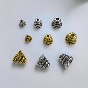 以下が含まれることがあります： 複雑なデザインが施された、銀と金の金属製の円錐形ビーズの詰め合わせ。ビーズはさまざまなサイズで、装飾的で華やかな外観をしています。