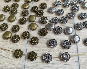 May include: A collection of small, round metal beads shaped like flowers, featuring a detailed petal design. The beads are finished in antique bronze and silver tones, with some beads strung on a thin wire and others scattered across the surface.