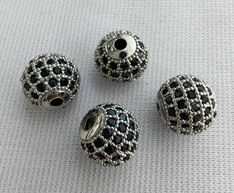 May include: Four silver round beads with a black diamond-like pattern. The beads have a hole in the centre for stringing.