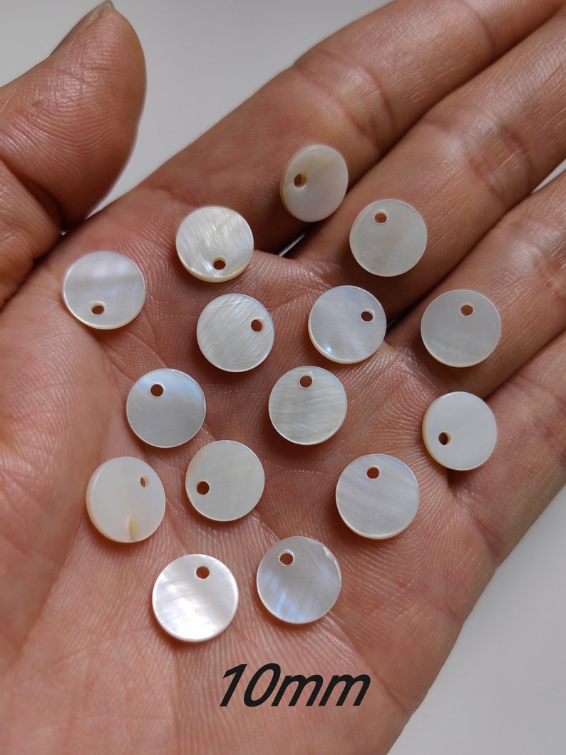 May include: A collection of 14 white, round, mother-of-pearl shell buttons with a hole in the center. The buttons are 10mm in diameter and are arranged in a loose pile on a hand.