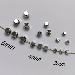 Może przedstawiać: Zestaw srebrnych metalowych koralików w kształcie sześcianu w trzech rozmiarach: 3 mm, 4 mm i 5 mm. Koraliki są ułożone na białym tle z przechodzącym przez nie złotym drutem.