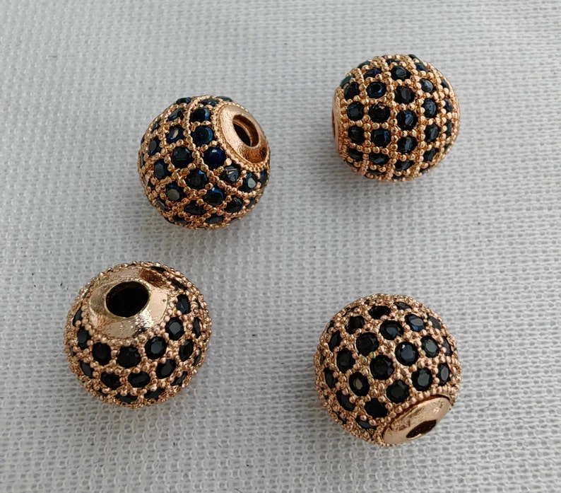 May include: Four gold-toned round beads with a black and gold geometric pattern. The beads are made of metal and have a shiny finish.
