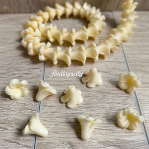 May include: A long string of cream white flower-shaped beads arranged in a coil, accompanied by several individual loose flower beads. Each bead features a central hole for threading. The text B# Cream White and findingscity is visible.