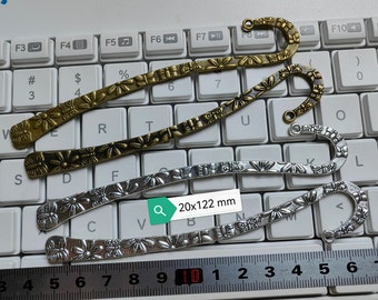 May include: Three decorative metal bookmarks with floral designs. The bookmarks are silver, gold, and bronze and have a hook at the top. The bookmarks are laid out on a keyboard with a ruler measuring 20x122 mm.