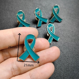 May include: A set of teal awareness ribbon pins with a metallic silver border. One pin is held in the foreground, showing dimensions of 26mm in height and 21mm in width. The other four pins are arranged in the background, each featuring the same design.