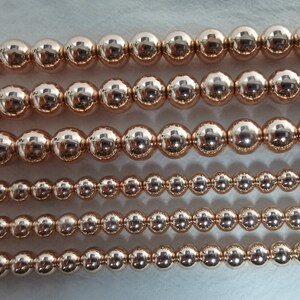 May include: Close-up of several strands of rose gold beads. The beads are perfectly round and have a high-shine finish, reflecting light. The sizes of the beads vary, creating visual interest. The beads are strung together, ready for jewelry making.