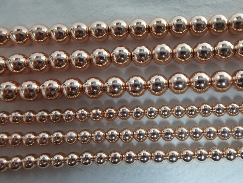May include: Close-up of several strands of rose gold beads. The beads are perfectly round and have a high-shine finish, reflecting light. The sizes of the beads vary, creating visual interest. The beads are strung together, ready for jewelry making.