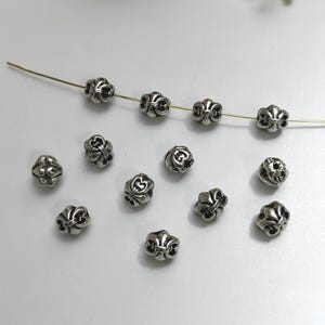 Può includere: Perline in metallo color argento con un design a fiore di giglio. Alcune perline sono infilate su un filo sottile, mentre altre sono sparse su una superficie bianca. Le perline hanno un aspetto dettagliato e ornato, adatte per la creazione di gioielli o lavori artigianali.