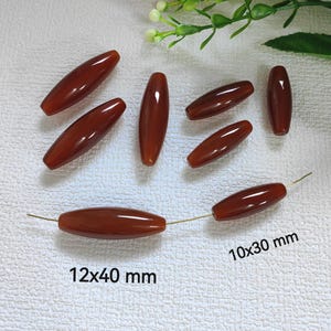 Può includere: Una collezione di perline lucide, di forma ovale, marrone rossiccio. Alcune perline sono infilate su un filo d'oro. L'immagine include il testo che indica le dimensioni: 12x40 mm e 10x30 mm. Adatto per la creazione di gioielli.