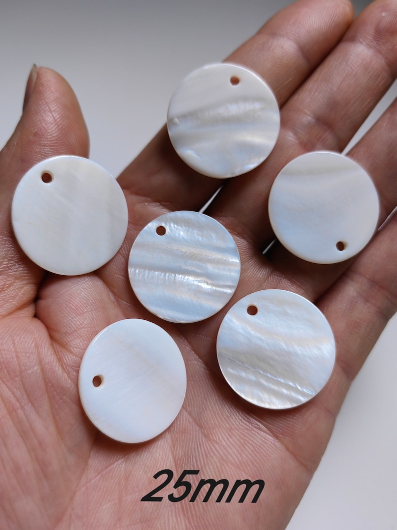 May include: Six white mother of pearl shell circles with a hole in the center. Each circle is approximately 25mm in diameter. The circles are arranged in a semi-circle on a hand.